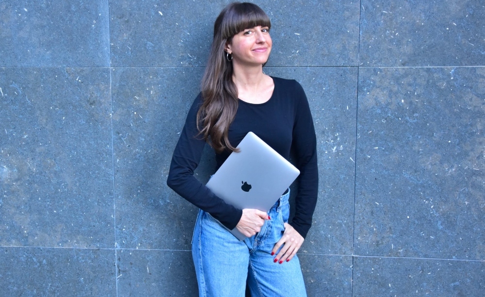A woman with brown hair posing for a business photo, holding a Macbook, wearing a black shirt and jeans